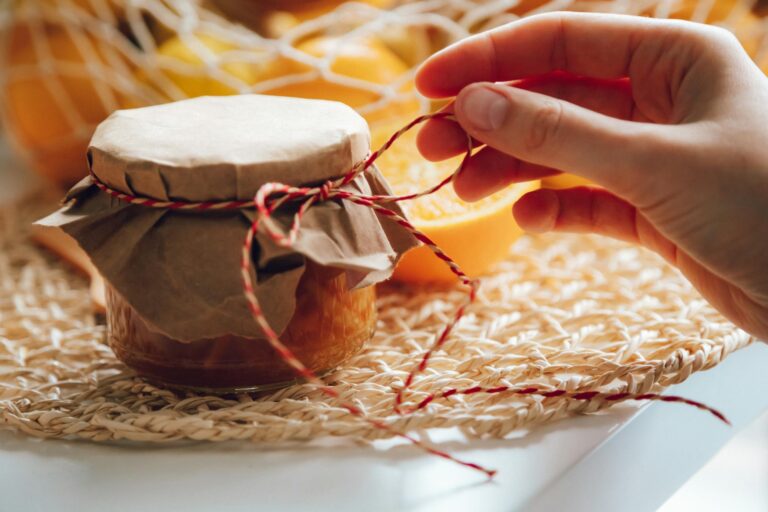 Homemade jam jar with hand tying string, surrounded by fresh oranges in warm lighting.